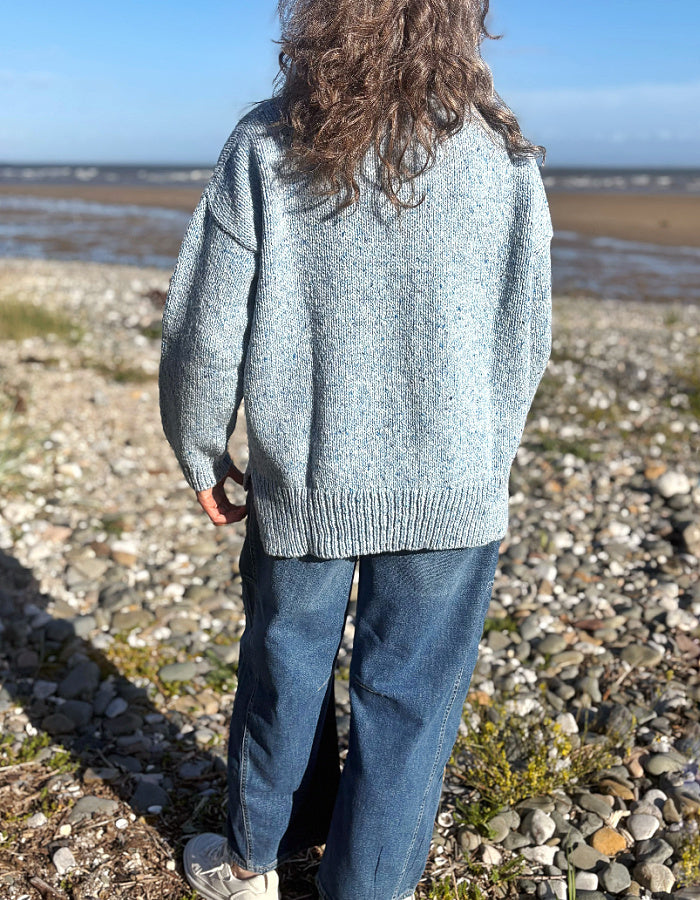 A woman wearing a light blue donegal tweed merino knit sweater and blue jeans stands on a rocky beach with water and sky in the background.