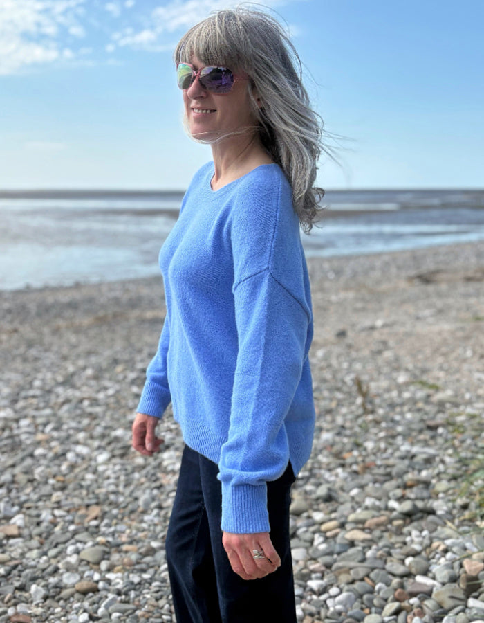 Woman in a blue sweater standing on a pebbly beach with a clear sky.