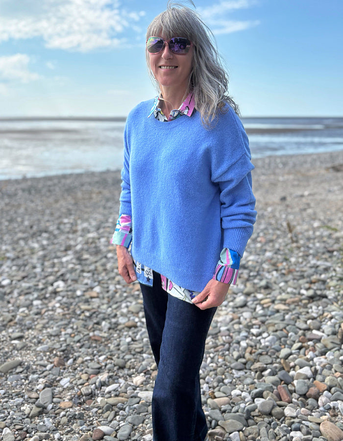 Woman in a blue sweater and sunglasses standing on a pebbly beach with ocean in the background
