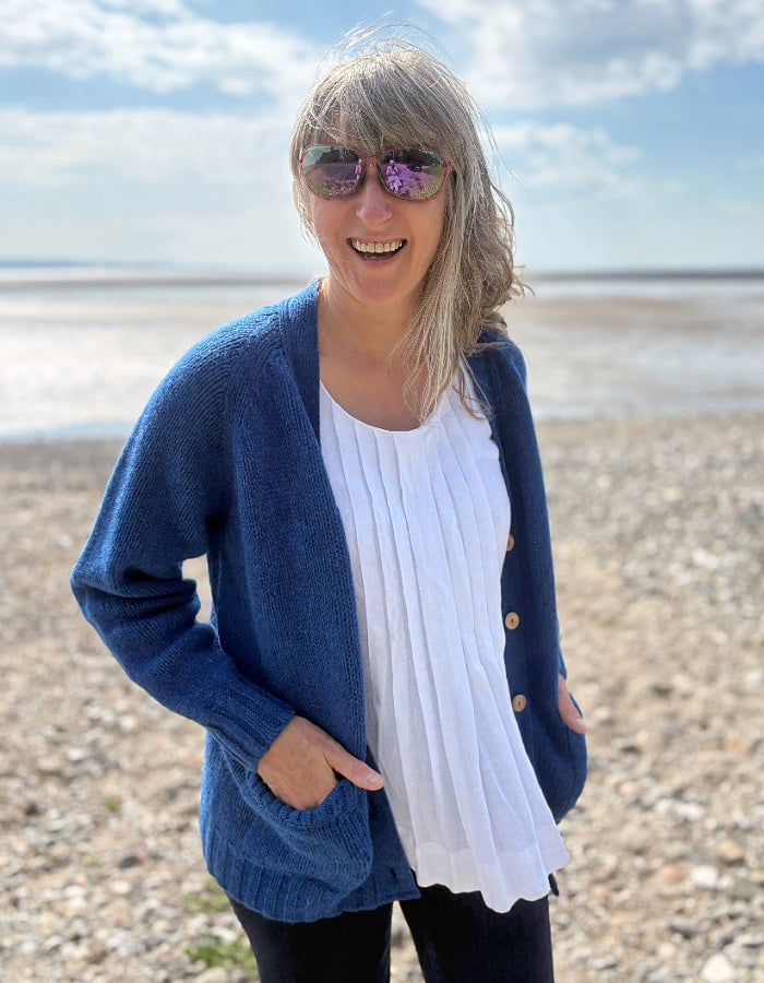 Woman wearing a wool blue cardigan on a pebbly beach with water and sky in the background