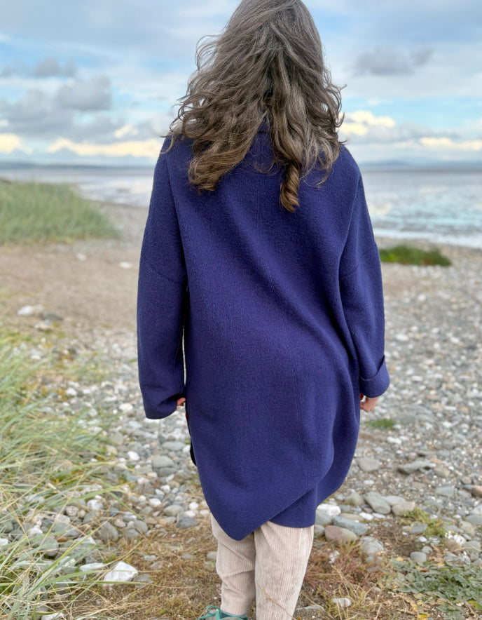 Person wearing a purple blue boiled wool coat standing on a beach with ocean and sky in the background