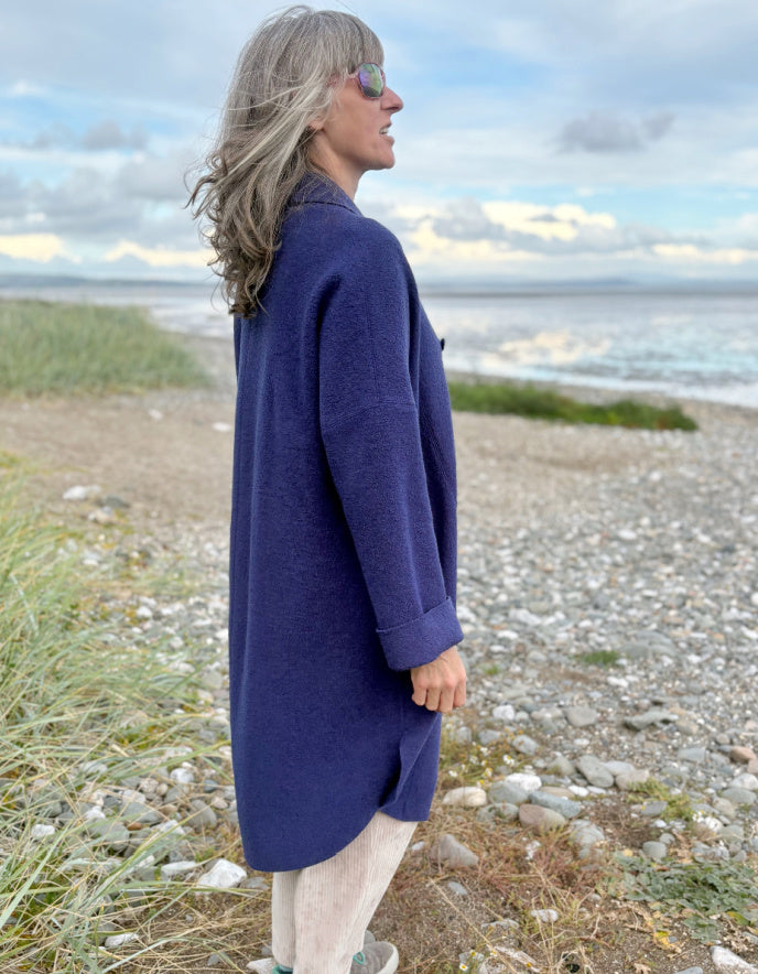Person wearing a purple blue boiled wool coat standing on a beach with ocean and sky in the background