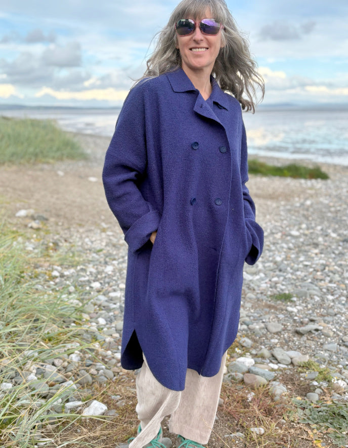 Person wearing a purple blue boiled wool coat standing on a beach with ocean and sky in the background