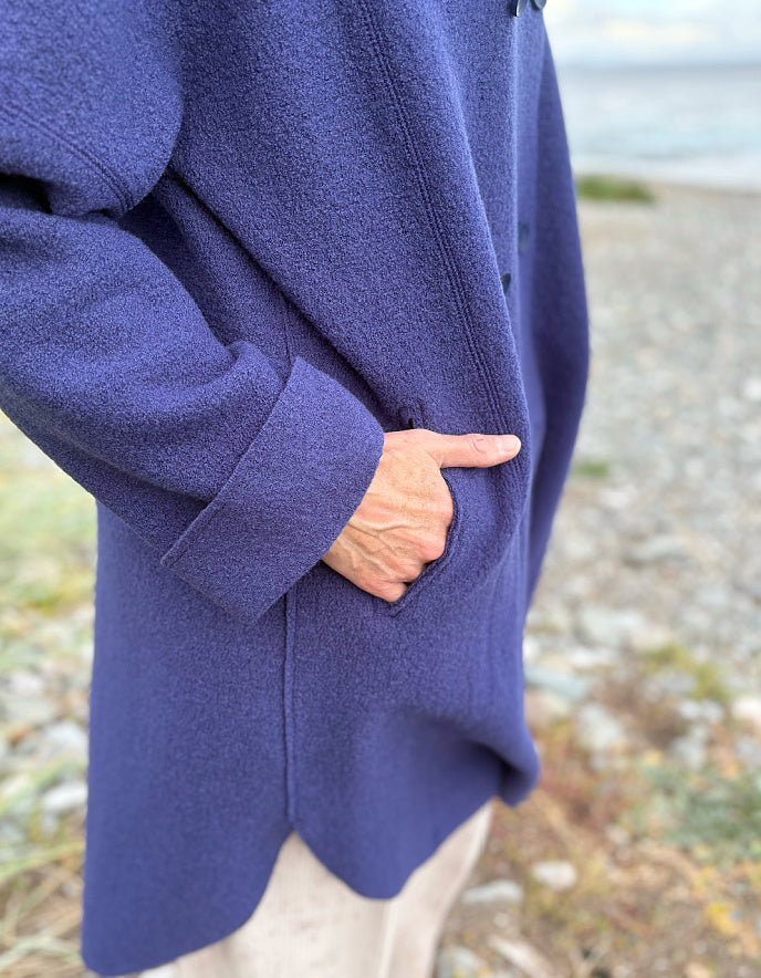 Person wearing a purple blue boiled wool coat standing on a beach with ocean and sky in the background
