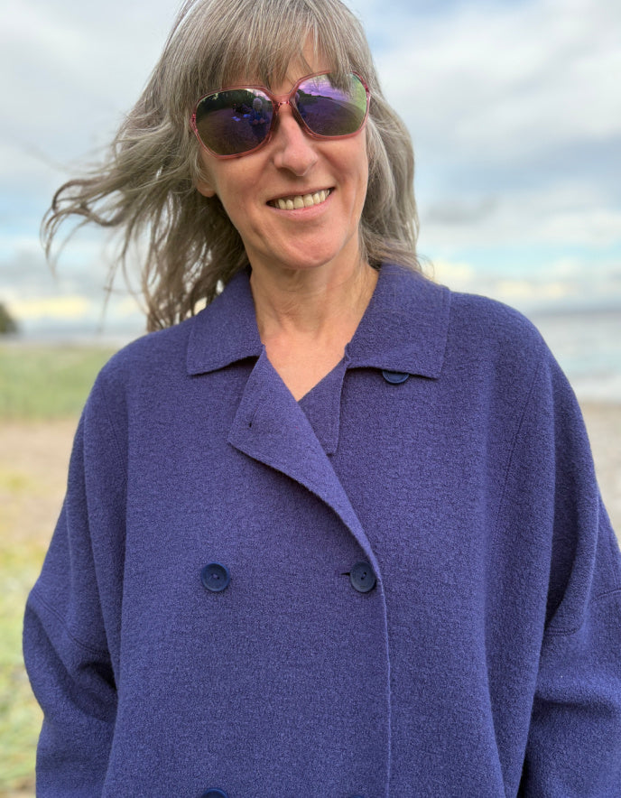 Person wearing a purple blue boiled wool coat standing on a beach with ocean and sky in the background