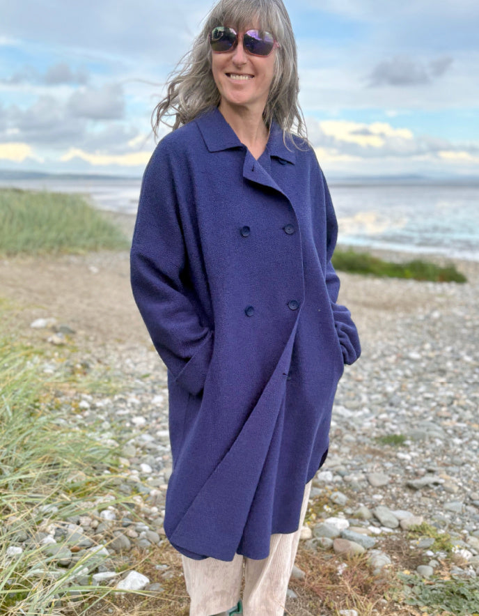 Person wearing a purple blue boiled wool coat standing on a beach with ocean and sky in the background