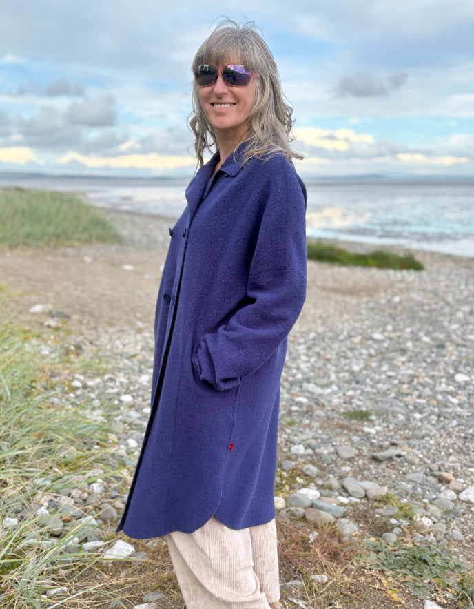 Person wearing a purple blue boiled wool coat standing on a beach with ocean and sky in the background