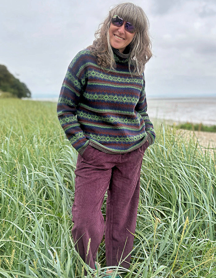 Person wearing a patterned fair Isle sweater and purple pants standing in a grassy field with a beach in the background.
