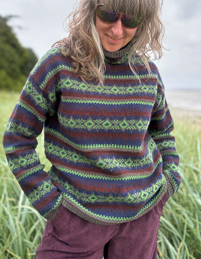 Person wearing a patterned fair Isle sweater and purple pants standing in a grassy field with a beach in the background.
