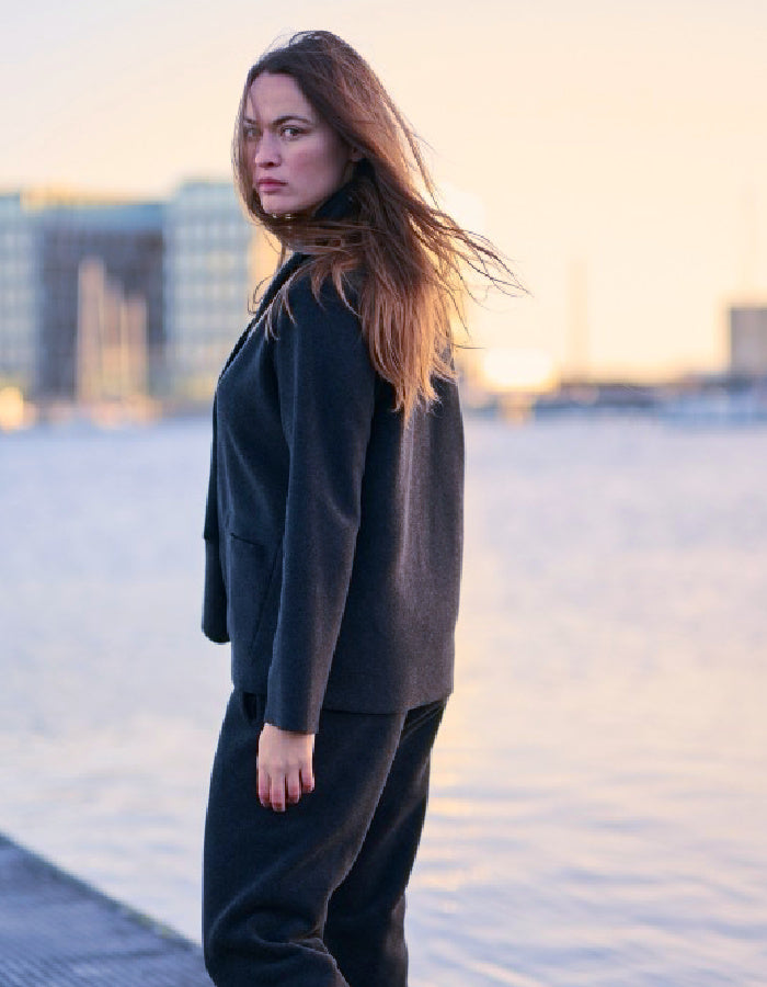 A woman with long light brown hair stands outdoors wearing a dark navy tailored jacket and matching dark trousers, with a softly blurred city waterfront in the background.