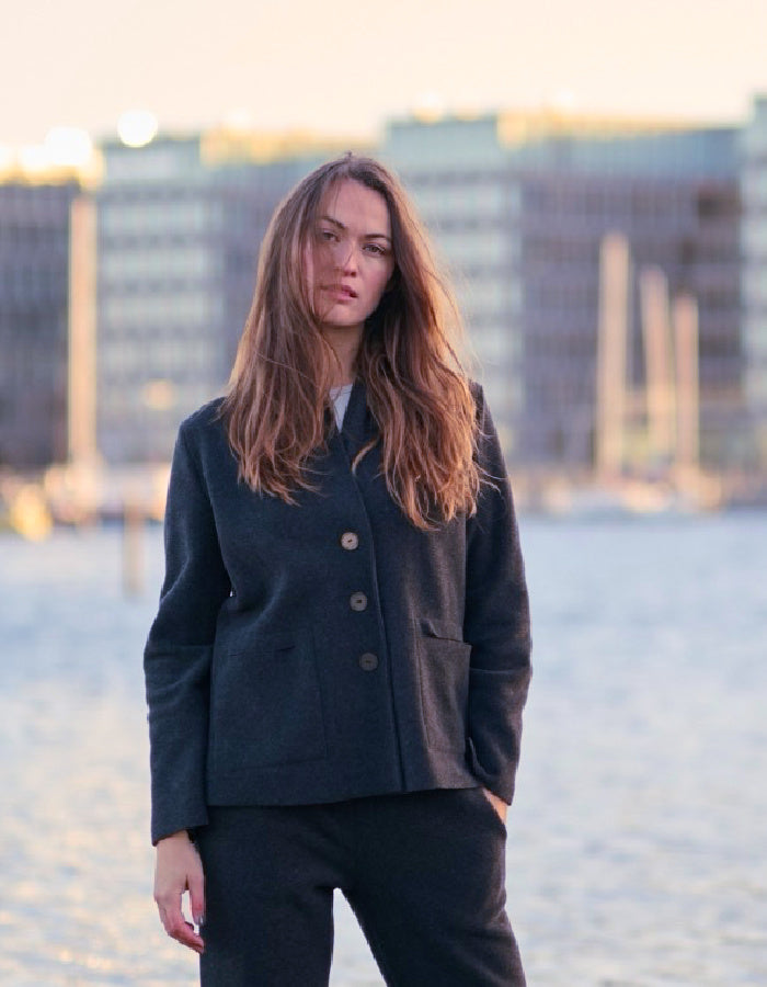 A woman with long light brown hair stands outdoors wearing a dark navy tailored jacket and matching dark trousers, with a softly blurred city waterfront in the background.