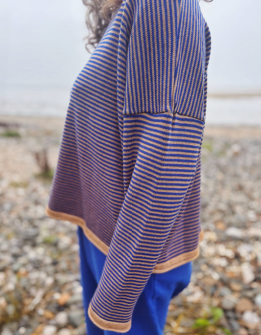 A woman wearing a blue beanie and a blue and camel striped knit sweater, standing on a pebble beach with water in the background.