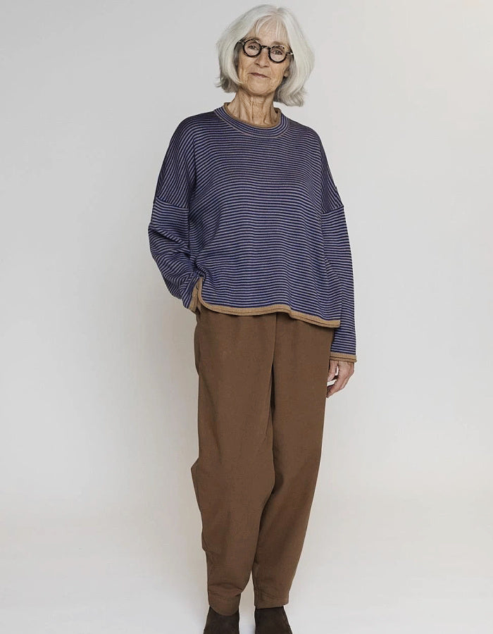 An older woman with short gray hair and glasses stands against a plain light gray studio background, wearing a loose navy and white striped long-sleeve top with brown pants and dark socks.