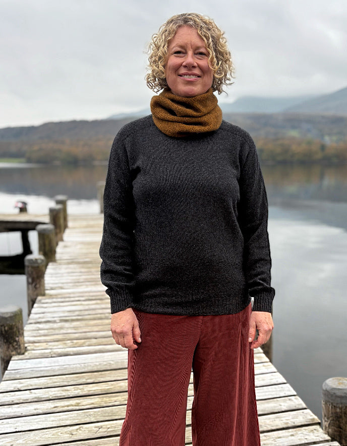 Woman standing on a wooden dock by a lake, wearing a dark sweater and rust-colored pants.
