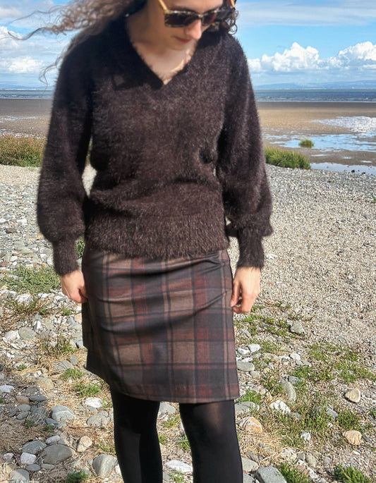 A person standing on a rocky beach wearing a dark brown fuzzy knit sweater, a brown and red plaid skirt, black tights, and sunglasses, with a body of water and cloudy sky in the background.