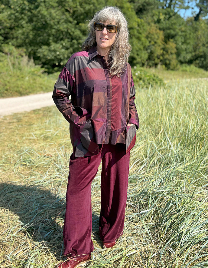 Woman wearing a check taffeta shirt and burgundy velvet pants standing in a field with trees in the background
