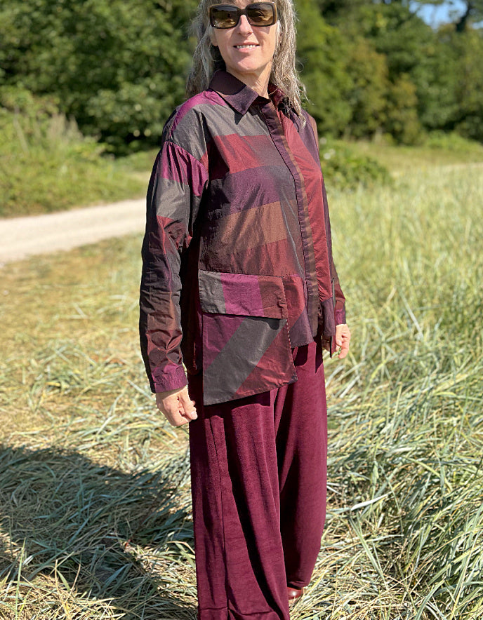 Woman wearing a check taffeta shirt and burgundy velvet pants standing in a field with trees in the background