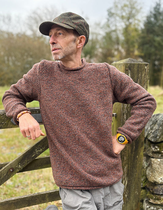 Man wearing a brown sweater and cap leaning against a wooden gate outdoors.