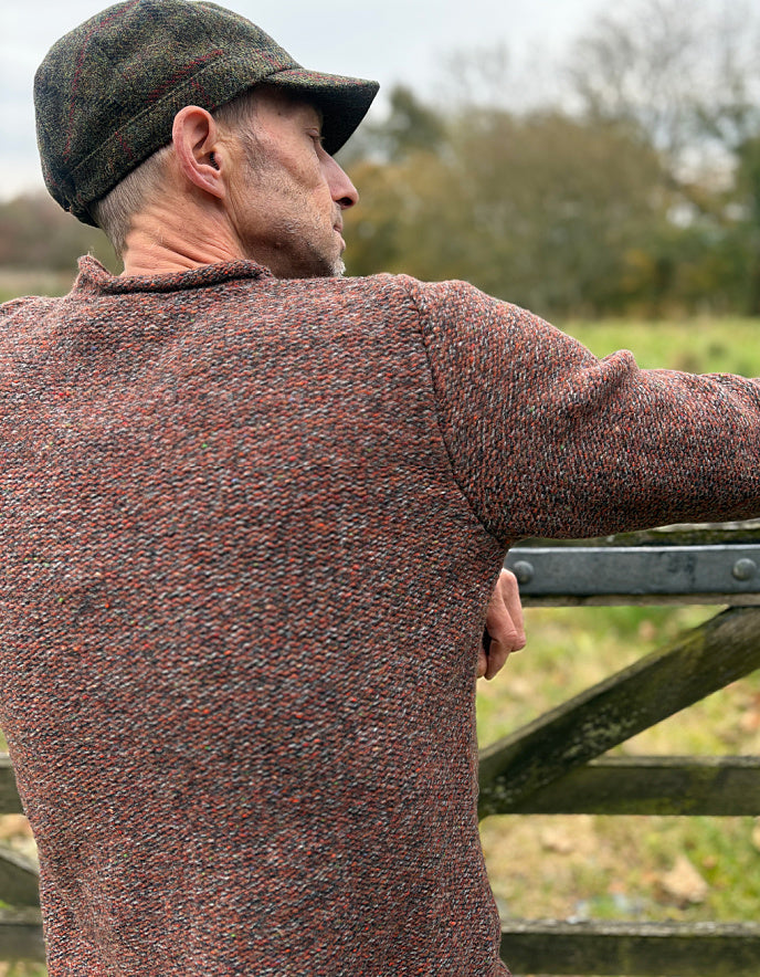 Man wearing a patterned sweater and cap, standing by a wooden gate in an outdoor setting.