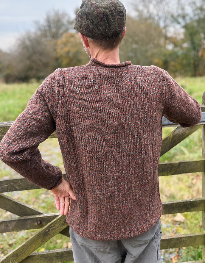 Man wearing a brown knitted sweater and cap standing by a wooden gate in a field.