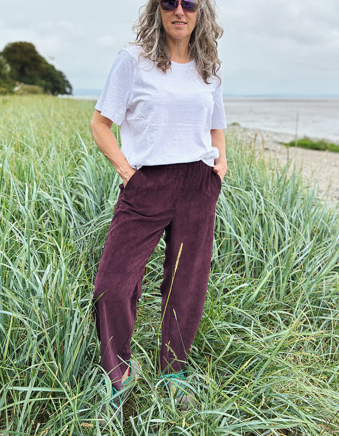 Woman standing in a grassy field wearing a white shirt and maroon wide leg corduroy trousers pants.
