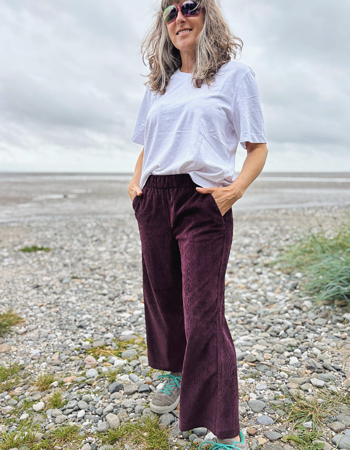 Woman standing on a pebbly beach wearing a white t-shirt and dark aubergine cord trousers pants.