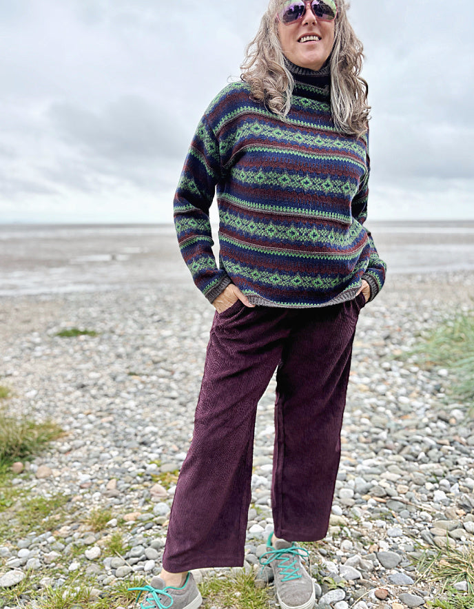Person wearing a patterned sweater and purple pants standing on a pebbly beach.