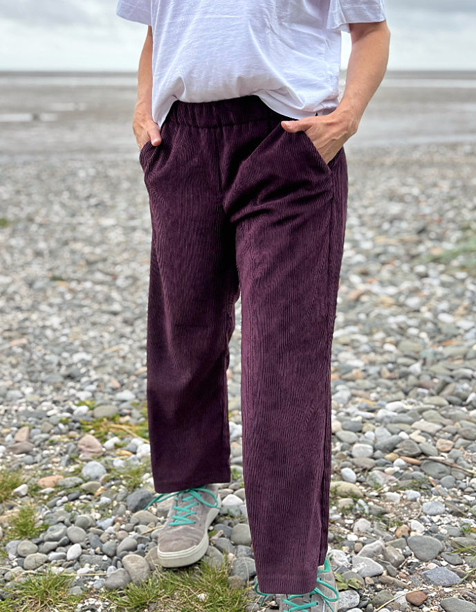 Person wearing purple cord pants and a white shirt standing on a pebbly beach.