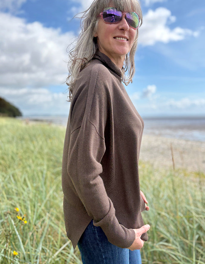 Person wearing a brown sweater with mock turtle neck standing in grass with a beach and blue sky in the background