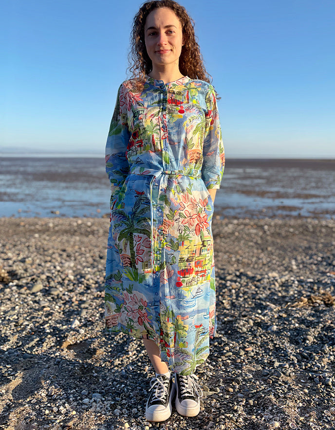 Woman in a colorful floral dress standing on a pebbly beach with a clear blue sky.