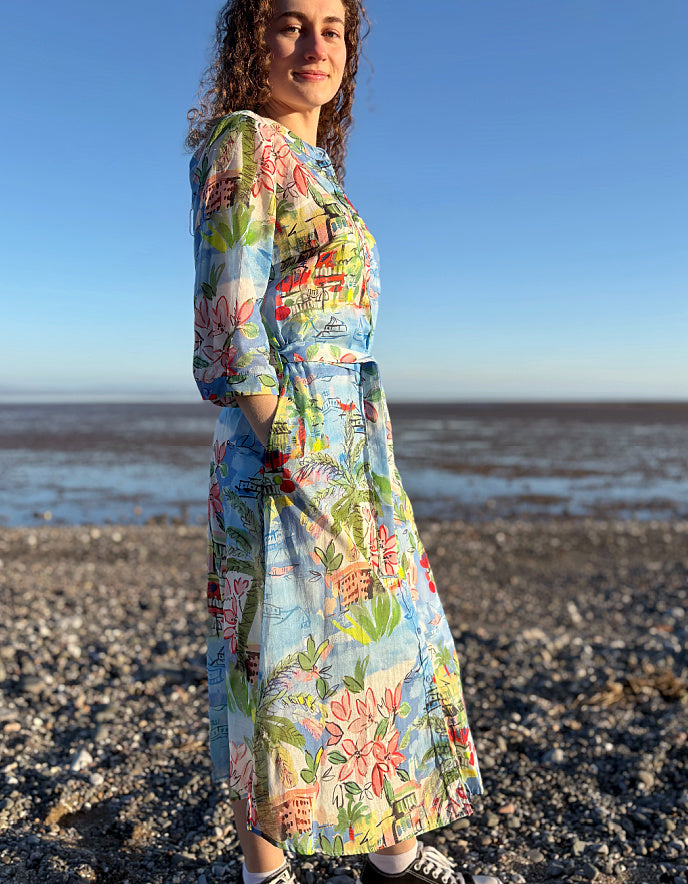 Woman wearing a colorful floral dress standing on a beach with a clear blue sky.