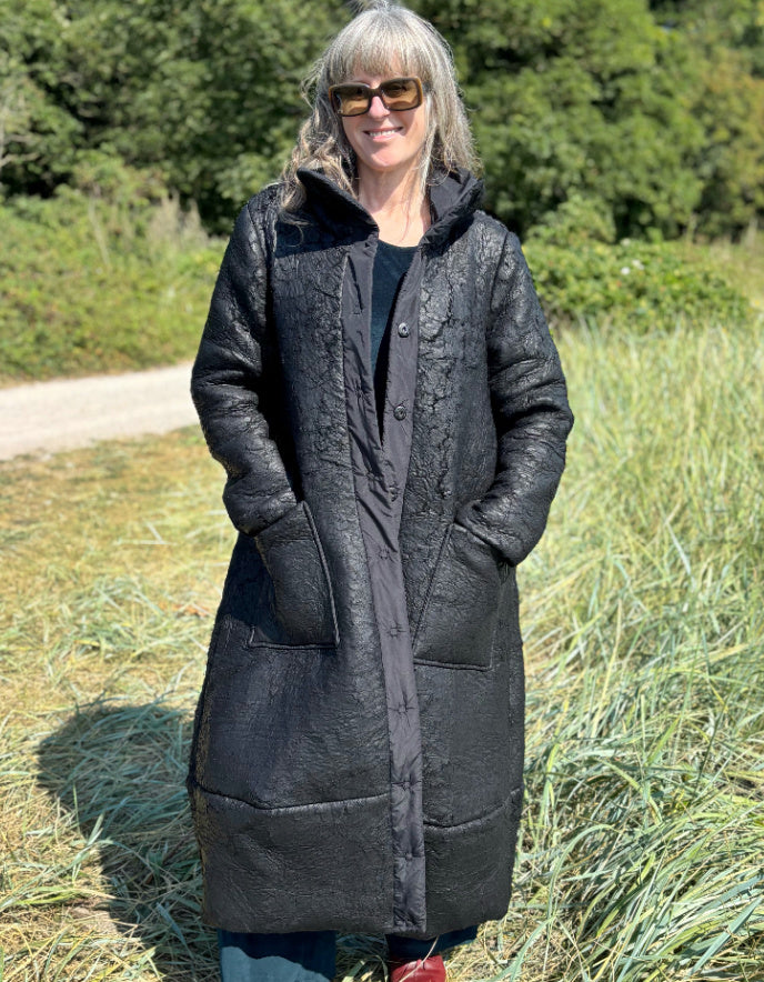 Person wearing a long dark coat standing in a grassy outdoor area.