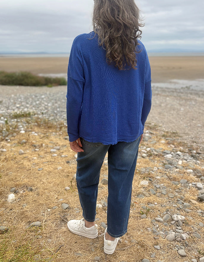 A person standing outdoors on a rocky, dry shoreline wearing a blue ribbed knit sweater, dark blue jeans, and white sneakers, photographed from behind.
