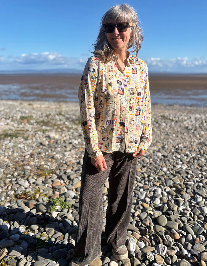 A woman with gray hair and sunglasses stands on a rocky beach wearing a beige button-up silky shirt covered in colourful illustrated print and dark brown cord wide leg pants, with water and blue sky in the background.
