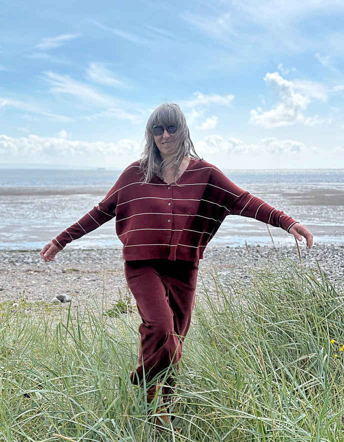 Person wearing a copper striped cardigan and pants standing in a grassy field with a beach in the background.