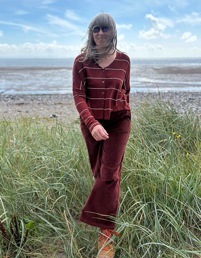 Person wearing a copper striped cardigan and pants standing in a grassy field with a beach in the background.