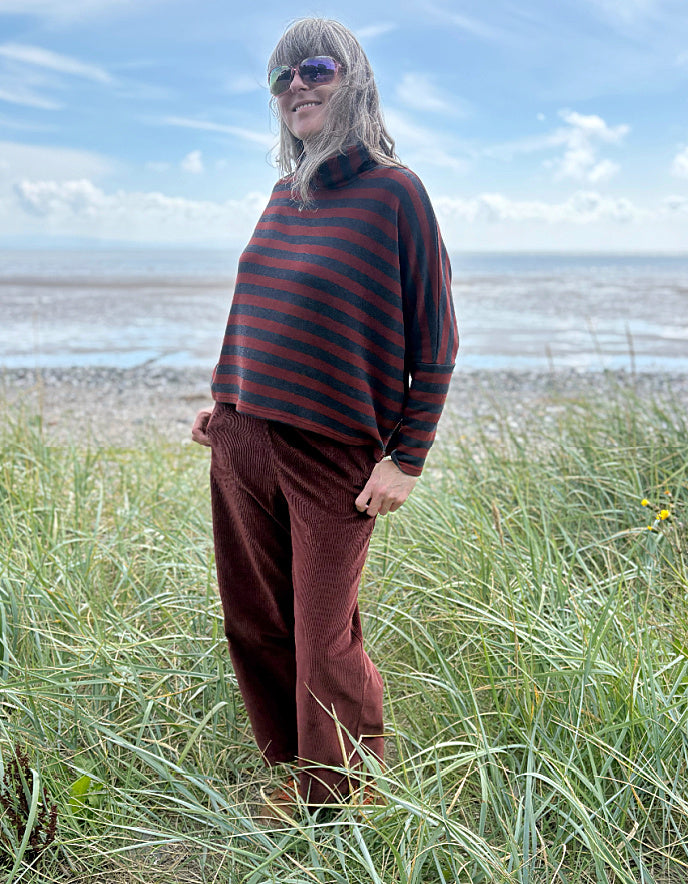 Person wearing a copper and charcoal striped turtle neck sweater with copper corduroy wide leg trousers and sunglasses on a beach