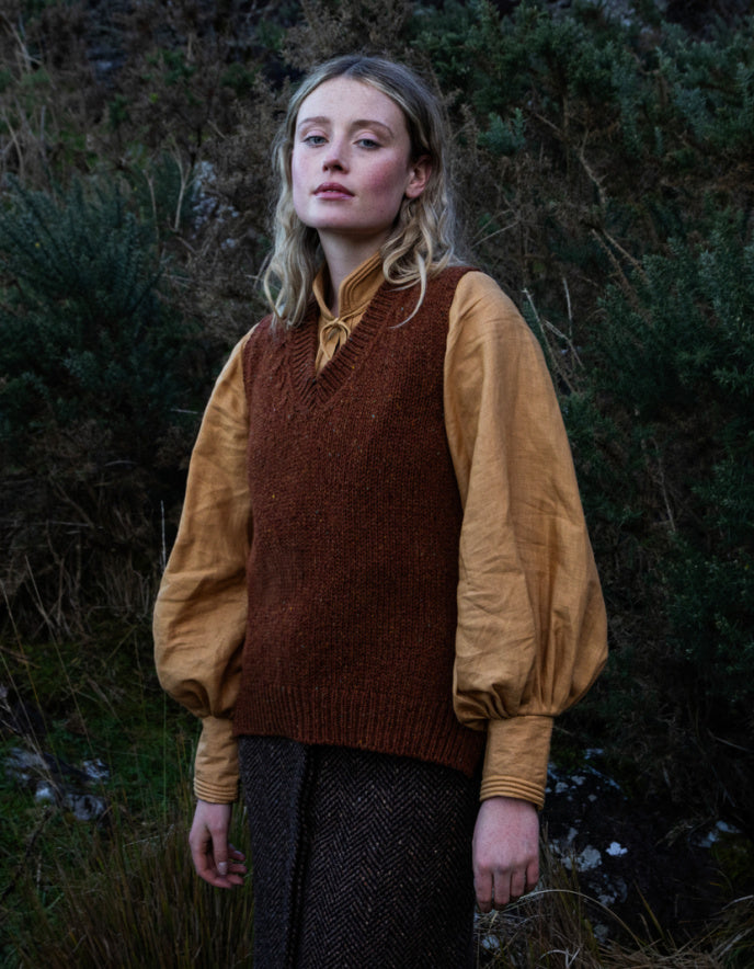 Woman wearing a copper brown donegal tweed tank top with a yellow shirt underneath, standing in a natural setting.