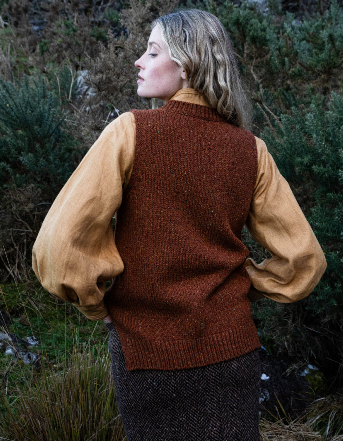 Woman wearing a copper brown donegal tweed tank top with a yellow shirt underneath, standing in a natural setting.