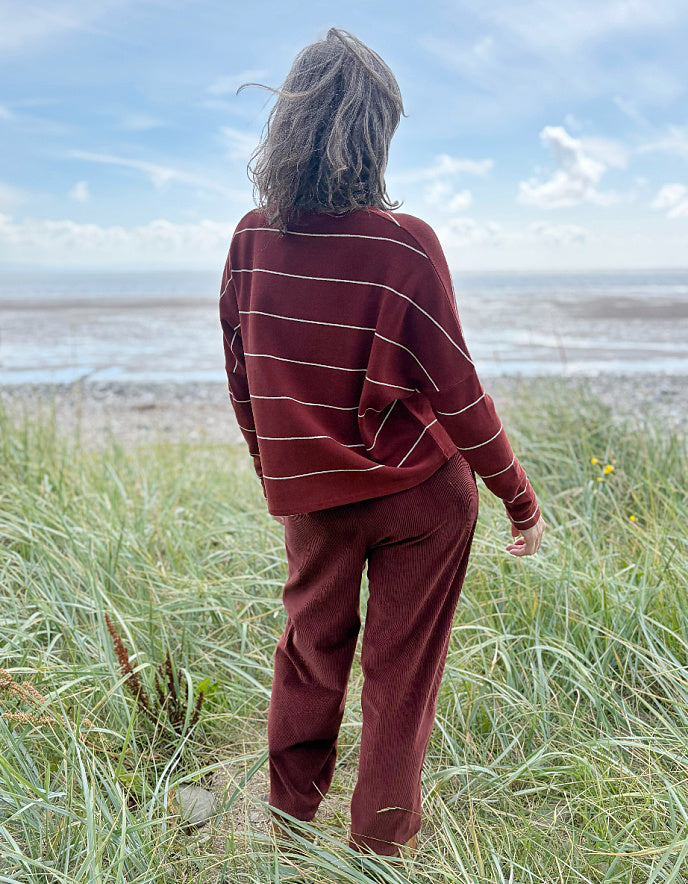 Person wearing a copper striped cardigan and pants standing in a grassy field with a beach in the background.