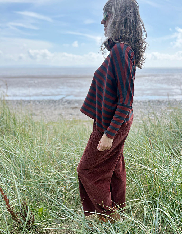 Person wearing a copper and charcoal striped turtle neck sweater with copper corduroy wide leg trousers and sunglasses on a beach