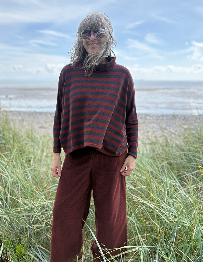 Person wearing a copper and charcoal striped turtle neck sweater with copper corduroy wide leg trousers and sunglasses on a beach