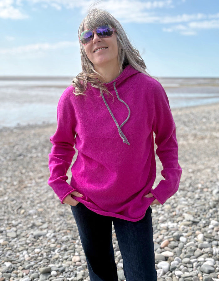 Person wearing a bright pink hoodie on a pebbly beach with a blue sky.