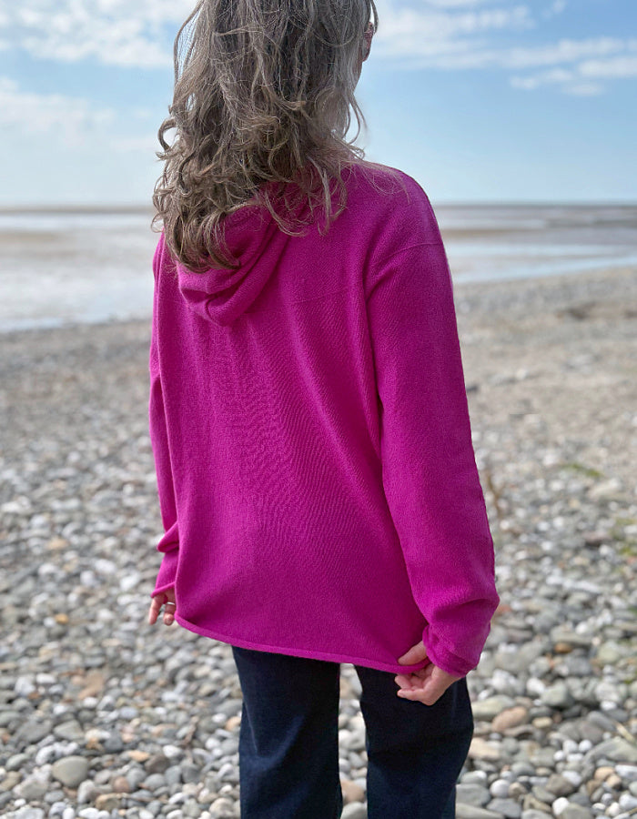 Person wearing a bright pink hoodie on a pebbly beach with a blue sky.