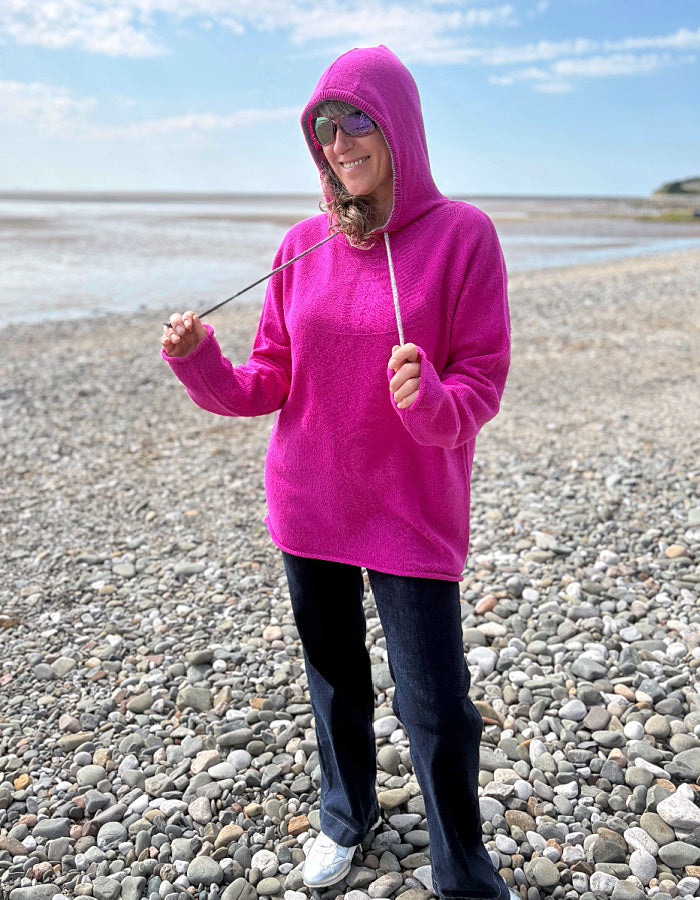 Person wearing a bright pink hoodie on a pebbly beach with clear skies.