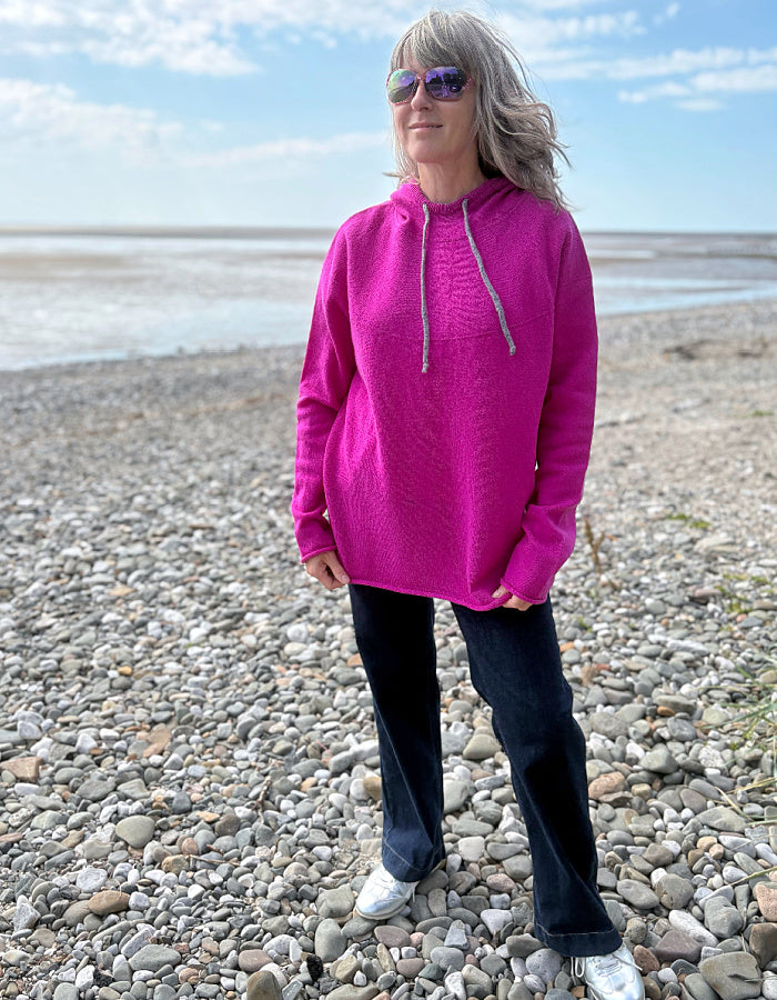 Person wearing a bright pink hoodie on a pebbly beach with a blue sky.