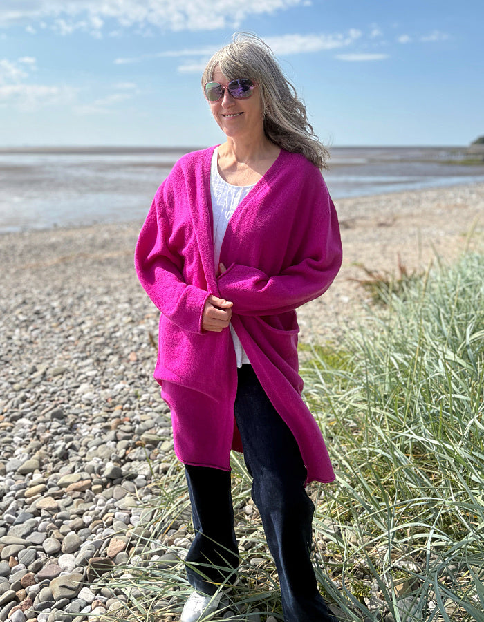 Person wearing a bright pink long wool cardigan standing on a pebbly beach with grass and water in the background.