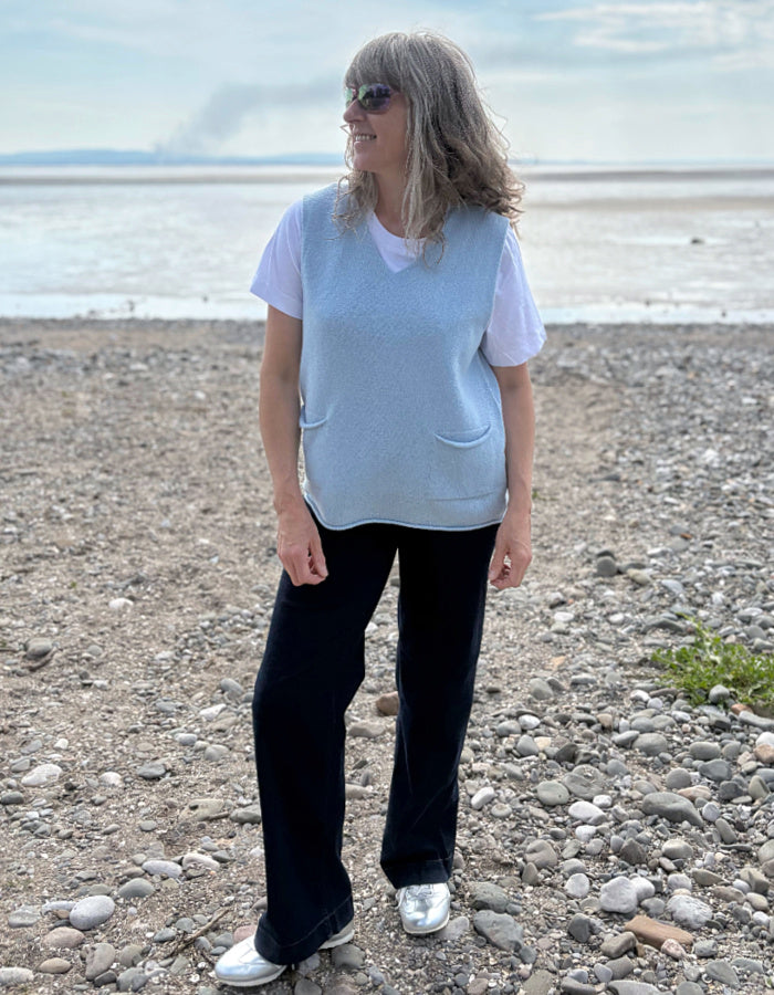 Woman standing on a pebbly beach wearing a light blue sleeveless top and dark wide leg jeans.