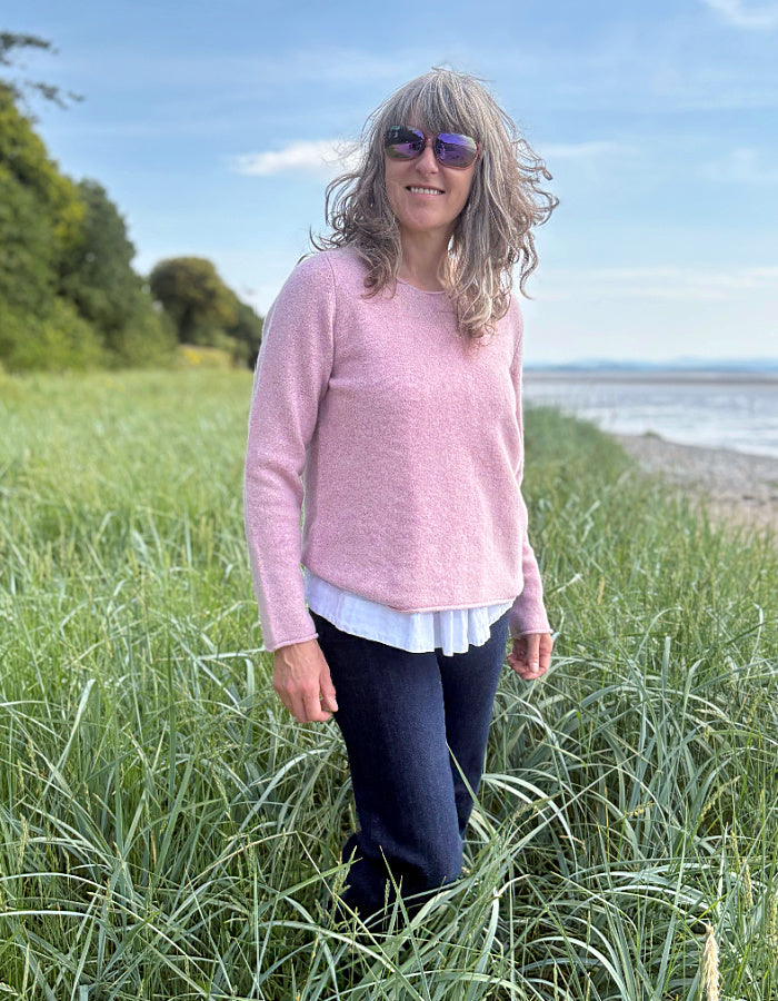 Woman in a pink wool sweater standing in tall grass by a coastal area