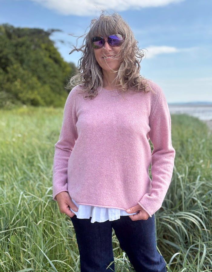 Woman in a pink wool sweater standing in tall grass by a coastal area